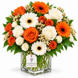 A vibrant floral arrangement in a clear square glass vase, featuring orange and white roses, orange and white gerbera daisies, and green foliage, with a 'FLOWRISTA' tag on the vase, set against a solid white background.