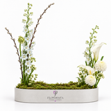 A floral arrangement in a white oval planter by FLOWRISTA, featuring white calla lilies, white dahlia, white flowers, green stems, moss, and tall bare twig branches on a white background.