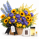 A floral gift basket arrangement in a woven basket with a black ribbon bow and tag reading FLOWRISTA, filled with yellow sunflowers, purple chrysanthemums, blue delphiniums, and yellow roses, shown with a boxed perfume and a small bottle on a white background.