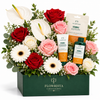 A floral gift box arrangement from Flowrista featuring red and pink roses, white and pink anthuriums, daisies, baby's breath, and three The Body Shop Vitamin C skincare products arranged in a dark green box.