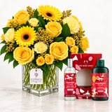 A bright yellow flower bouquet in a clear square glass vase with a FLOWRISTA tag, displayed beside two The Body Shop Cranberry Crush bath and body products on a white marble surface against a white background.