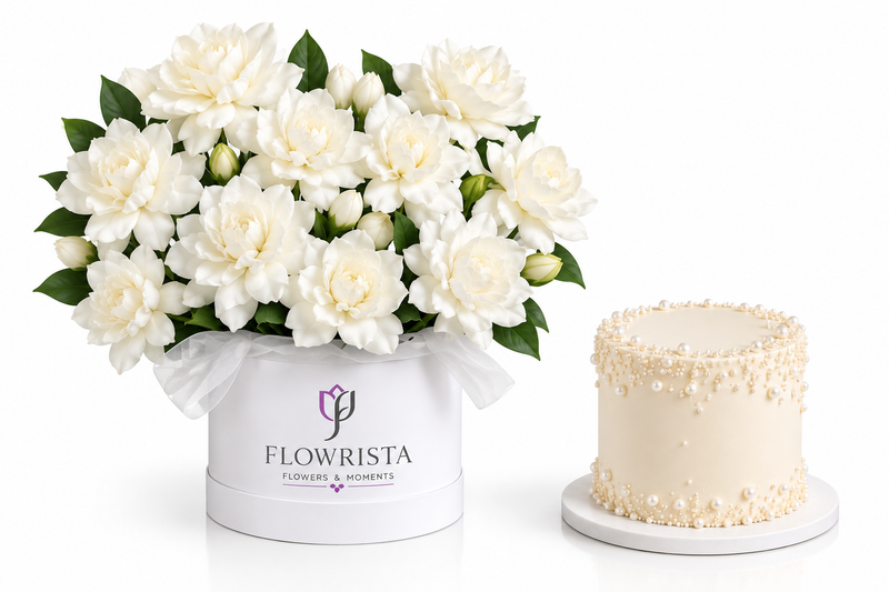 A white floral box filled with white peonies and green leaves, next to a small round cream-colored cake with decorative drizzle, on a clean white background. The box is branded FLOWRISTA.