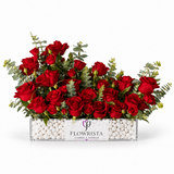 A rectangular clear acrylic flower box filled with white decorative pebbles and a white label reading FLOWRISTA, filled with a dense arrangement of red roses and green eucalyptus stems, photographed on a clean white background.