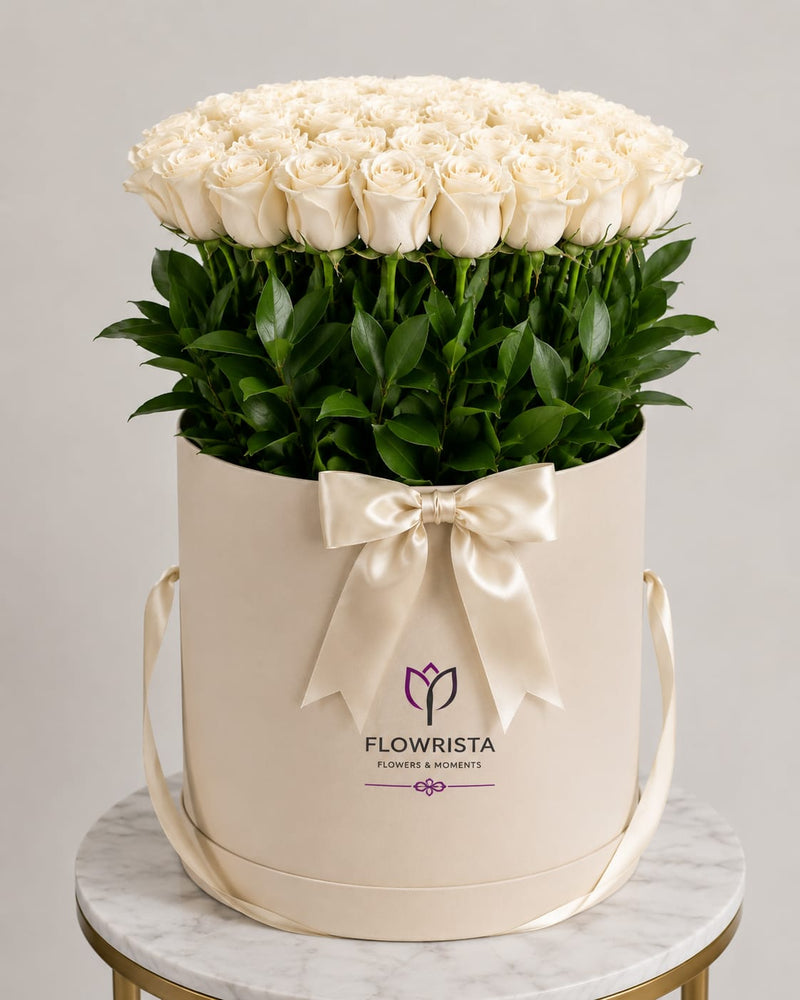 A large round cream-colored gift box filled with a dense arrangement of white roses and green stems, tied with a cream satin bow. The box is branded 'FLOWRISTA' and sits on a marble-topped table against a light gray background.