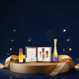 A luxury fragrance gift set displayed on a round gold pedestal against a dark navy blue background with gold bokeh lights and ribbon accents. The set includes a small yellow perfume bottle with a red cap, an open silver box containing three small cylindrical bottles, and a tall iridescent bottle with a gold cap and purple base.