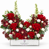 A floral arrangement in a rectangular clear planter filled with white pebbles, featuring red roses, white roses, burgundy orchids, green foliage, and red berry clusters on a white background. The planter is branded with the Flowrista logo.