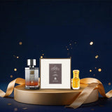 A luxury fragrance gift set featuring a dark glass bottle with a silver cap, a small yellow perfume bottle, and a textured cream-colored box, arranged on a round gold pedestal with a gold ribbon against a dark blue background with gold confetti.