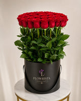 A large bouquet of 60 fresh red roses with green stems and leaves, presented in a black cylindrical container with the brand name 'FLOWRISTA' printed on the front, set on a marble-topped table against a neutral beige background.