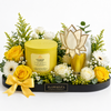A curated floral gift arrangement on a black tray from Flowrista, featuring a yellow Kitchen Lemon scented candle tin, a gold-and-white lotus-shaped glass diffuser, yellow and white roses, daisies, green foliage, and a yellow ribbon bow, photographed on a white background.