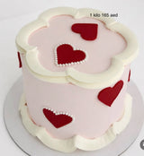 A round Valentine's Day cake decorated with pale pink icing, white piped frosting borders, and red heart shapes accented with small white pearl-like dots. Text in the upper right reads '1 kilo 165 aed'.