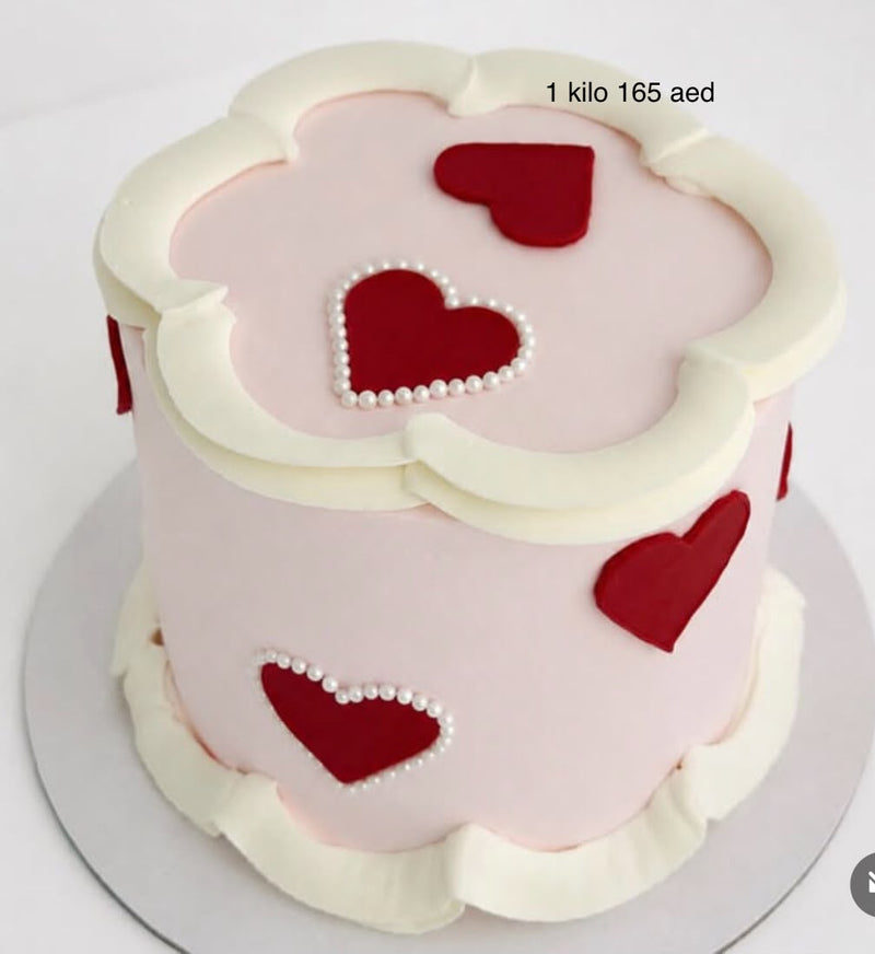 A round Valentine's Day cake decorated with pale pink icing, white piped frosting borders, and red heart shapes accented with small white pearl-like dots. Text in the upper right reads '1 kilo 165 aed'.