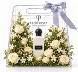A clear acrylic gift box with a handle, tied with a lavender ribbon, containing a bottle of Flowrista 'White & Bright Vanilla' perfume surrounded by cream roses, white flowers, and green foliage on a white background.