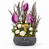 A floral arrangement in a gray stone-style pot by Flowrista, featuring purple anthuriums, white roses, green foliage, and a small gold-and-black candle, photographed on a clean white background.