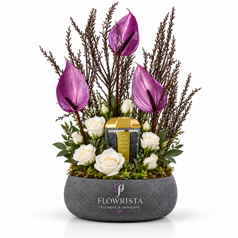 A floral arrangement in a gray stone-style pot by Flowrista, featuring purple anthuriums, white roses, green foliage, and a small gold-and-black candle, photographed on a clean white background.