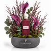 A floral arrangement in a round gray stone planter featuring a central textured burgundy bottle with a white cap and a label reading 'PURPLE BEAR ESSENTIALS,' surrounded by purple calla lilies, green eucalyptus, and pinkish filler stems on a white background. The planter is branded FLOWRISTA FLOWERS & MOMENTS.