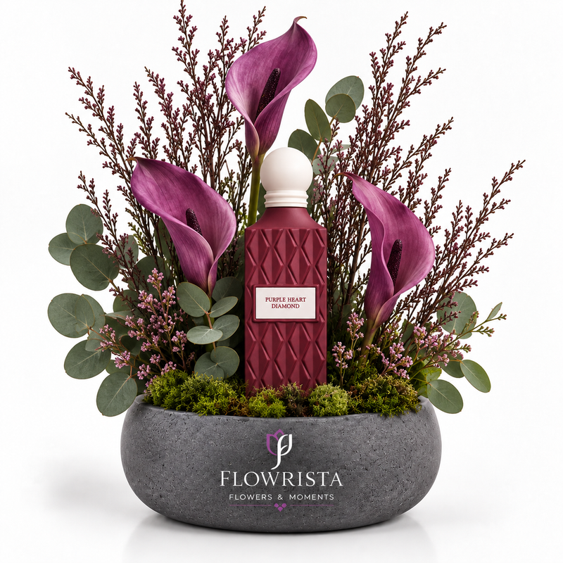 A floral arrangement in a round gray stone planter featuring a central textured burgundy bottle with a white cap and a label reading 'PURPLE BEAR ESSENTIALS,' surrounded by purple calla lilies, green eucalyptus, and pinkish filler stems on a white background. The planter is branded FLOWRISTA FLOWERS & MOMENTS.
