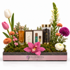 A floral gift basket arrangement by Flowrista featuring various perfume bottles, a candle, and a jar of essential oils nestled in green moss and surrounded by colorful flowers, including tulips, an orchid, and chrysanthemums, in a pink and dark gray wooden tray.