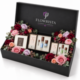 A luxury gift set from Flowrista Flowers & Moments in an open black box, decorated with pink, red, and purple roses and green eucalyptus leaves, containing skincare and cosmetic products.