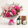 A large pink flower bouquet wrapped in white tissue paper with a pink ribbon, displayed alongside The Body Shop skincare products including a shower gel bottle, a hand cream tube, and a body butter tin, with their branded gift box in the background.