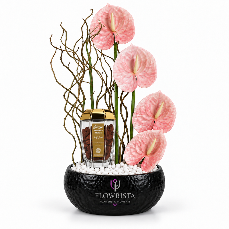 A floral arrangement in a black textured bowl filled with white pebbles, featuring four pink anthuriums, bare decorative branches, and a small clear jar of coffee with gold lids. The arrangement is set against a plain white background with the brand name 'FLOWRISTA' visible on the front of the bowl.