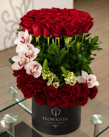 A large floral arrangement of red roses, white orchids, and green foliage in a black cylindrical container branded FLOWRISTA, placed on a glass table in an indoor setting.