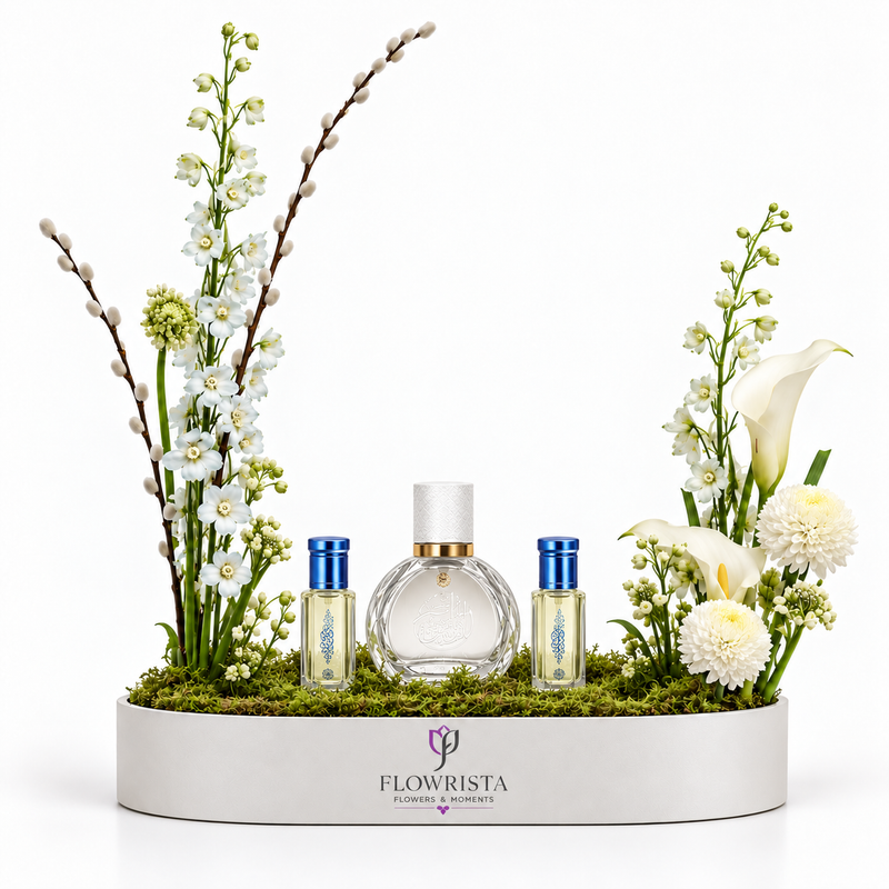 FLOWRISTA perfume gift set arranged in a white oval planter with green moss and white flowers. The set includes a large clear glass perfume bottle with a white cap and gold collar, two small cylindrical bottles with blue caps, and floral arrangements with white calla lilies, white flowers, and green stems.