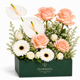 A floral arrangement in a dark green rectangular box from Flowrista, featuring peach roses, white anthuriums, white gerbera daisies, small white roses, and green filler flowers on a clean white background.