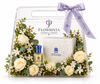 A clear acrylic gift basket from Flowrista, decorated with white roses, baby's breath, and green eucalyptus leaves, tied with a lavender ribbon. Inside are a small amber perfume bottle with a blue cap, a white patterned box, and a floral arrangement.