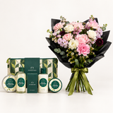 A curated gift set from The Bath Shop featuring a large bouquet of pink, white, and burgundy flowers wrapped in black paper, alongside a matching bath and body set including a gift box, shower gel, body wash, hand cream, and a bath bomb tin.