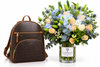 A brown patterned backpack with tan leather trim stands next to a large, vibrant flower bouquet in a clear glass vase labeled FLOWWRISTA, set against a clean white background.