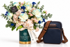 A floral arrangement in a Flowrista-branded green and gold vase beside a navy blue crossbody bag with brown straps, photographed on a clean white background.