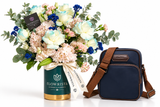 A floral arrangement in a Flowrista-branded green and gold vase beside a navy blue crossbody bag with brown straps, photographed on a clean white background.
