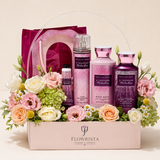 A Flowrista gift basket filled with pink and purple personal care products, including body wash, body lotion, body spray, and a body mist, arranged with pink, peach, and white flowers in a light pink box with a clear plastic handle.