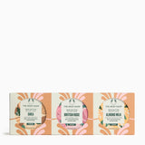 Three round body butter bars in a matching cardboard gift set from The Body Shop. The packaging features a peach and cream floral design with circular cutouts showing the colorful labels of Shea, British Rose, and Almond Milk scents.
