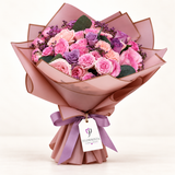 A vibrant bouquet of pink and purple roses wrapped in dusty pink paper with a purple ribbon and a 'FLOWRISTA' tag, set against a clean white background.