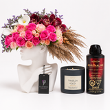 A curated gift set featuring a vibrant pink and red floral arrangement in a white female bust vase, a black candle labeled Vanilla Oud, a black body spray can labeled Adrenaline, and a black tag from Flowrista.