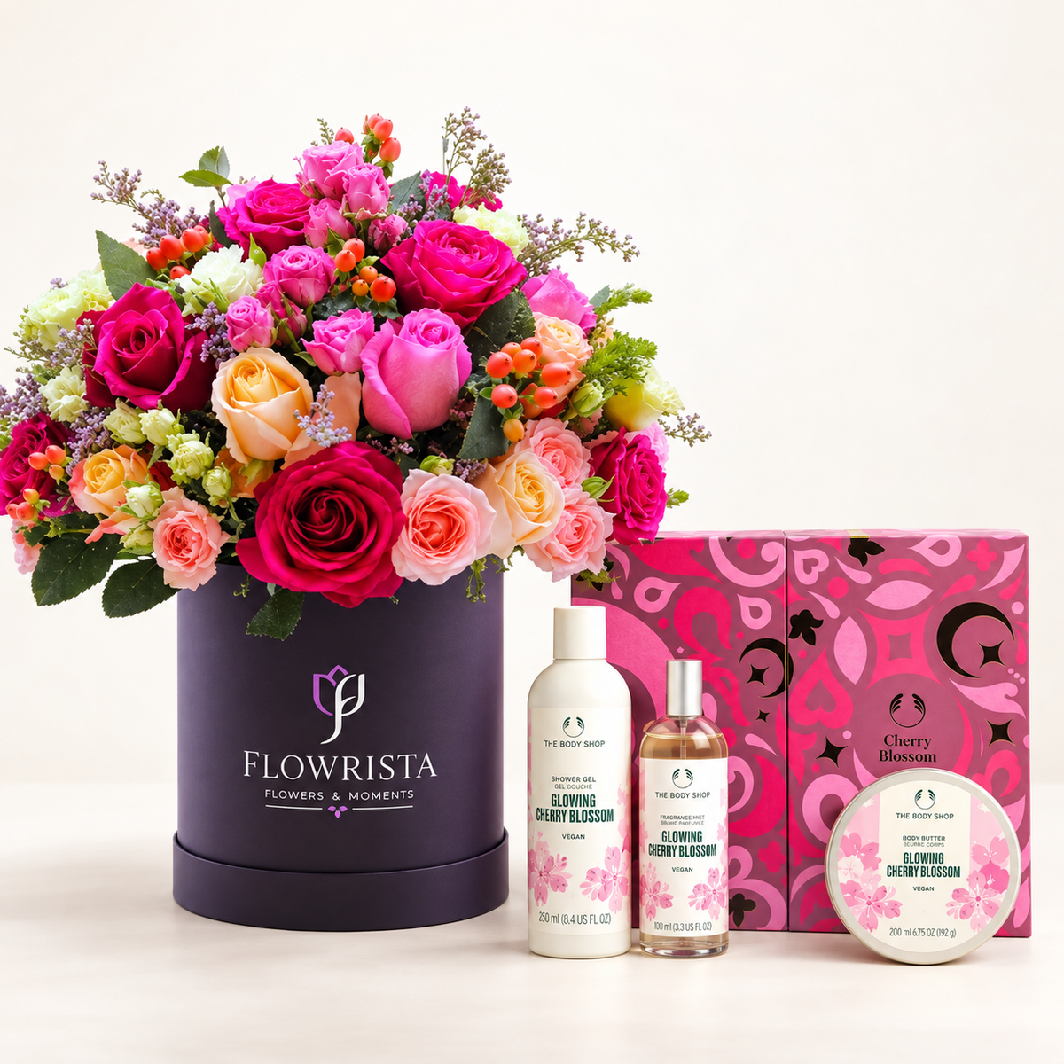 A curated gift set featuring a large bouquet of pink, peach, and orange roses in a dark purple round container labeled 'FLOWRISTA', alongside three personal care products from The Body Shop: a glowing cherry blossom body wash, a glowing cherry blossom body lotion, and a glowing cherry blossom body butter, with a pink patterned gift box behind them.