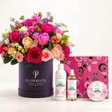 A curated gift set featuring a large bouquet of pink, peach, and orange roses in a dark purple round container labeled 'FLOWRISTA', alongside three personal care products from The Body Shop: a glowing cherry blossom body wash, a glowing cherry blossom body lotion, and a glowing cherry blossom body butter, with a pink patterned gift box behind them.
