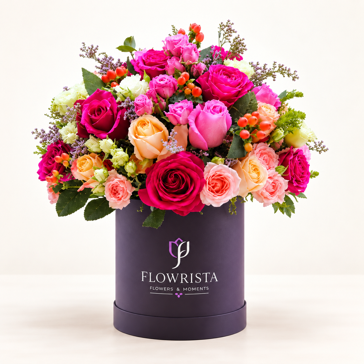 A vibrant floral arrangement of pink, magenta, peach, and orange roses with green foliage, presented in a dark purple cylindrical box printed with the brand name 'FLOWRISTA'.