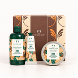 The Body Shop Shea body care gift set featuring a shower cream bottle, a hand balms tube, and a body butter tin, displayed in front of their brown and beige patterned gift box on a white background.