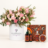 A curated gift set featuring a large pink and white flower arrangement in a white cylindrical container labeled 'FLOWRISTA', alongside matching home fragrance products including two dark bottles, a clear spray bottle, and a round tin with patterned labels, all arranged on a clean white background.