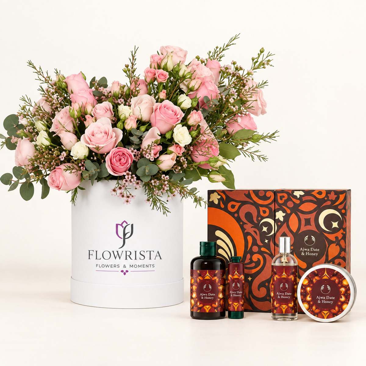 A curated gift set featuring a large pink and white flower arrangement in a white cylindrical container labeled 'FLOWRISTA', alongside matching home fragrance products including two dark bottles, a clear spray bottle, and a round tin with patterned labels, all arranged on a clean white background.