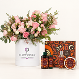 A curated gift set featuring a large pink and white flower arrangement in a white cylindrical container labeled 'FLOWRISTA', alongside matching home fragrance products including two dark bottles, a clear spray bottle, and a round tin with patterned labels, all arranged on a clean white background.