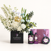 A curated gift set featuring a large floral arrangement in a black Flowerista box, alongside matching personal care products from The Body Shop including a shower gel bottle, hand cream tube, and body butter tin with a purple box behind them.