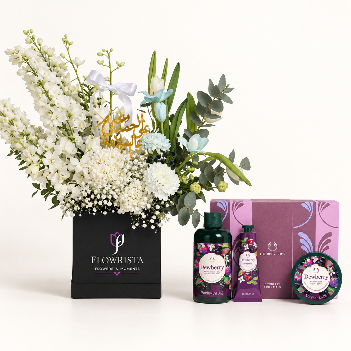 A curated gift set featuring a large floral arrangement in a black Flowerista box, alongside matching personal care products from The Body Shop including a shower gel bottle, hand cream tube, and body butter tin with a purple box behind them.