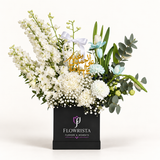 A floral arrangement in a black square box from Flowrista, featuring white and light blue flowers, green foliage, a white ribbon, and a gold Arabic script topper, photographed on a clean white background.