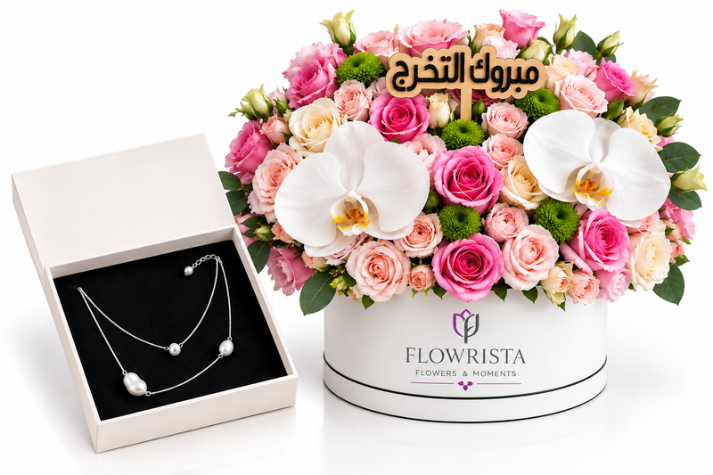A floral gift arrangement in a round white Flowerista box next to an open cream jewelry box containing a silver necklace with pearl-like beads. The bouquet features pink and peach roses, white orchids, green foliage, and a small wooden card with Arabic text.