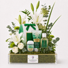 A floral gift basket arrangement from Flowrista, featuring white lilies, green roses, eucalyptus, and botanical products including a green soap bar, a green body wash tube, and a green shampoo bottle in a clear rectangular moss-lined basket with a white Flowrista label.