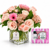 A floral arrangement in a clear square glass vase with pink and blush roses, pink gerbera daisies, and green foliage, displayed beside a pink and white The Body Shop Body Butter gift box on a white background.