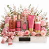 A curated gift basket from Flowrista featuring multiple pink and rose-themed personal care products arranged in a clear tray filled with white pebbles, decorated with pink roses, eucalyptus, and a pink satin bow. Visible products include a rose body wash, rose body mist, rose body lotion, rose hand cream, and a rose body butter tube.